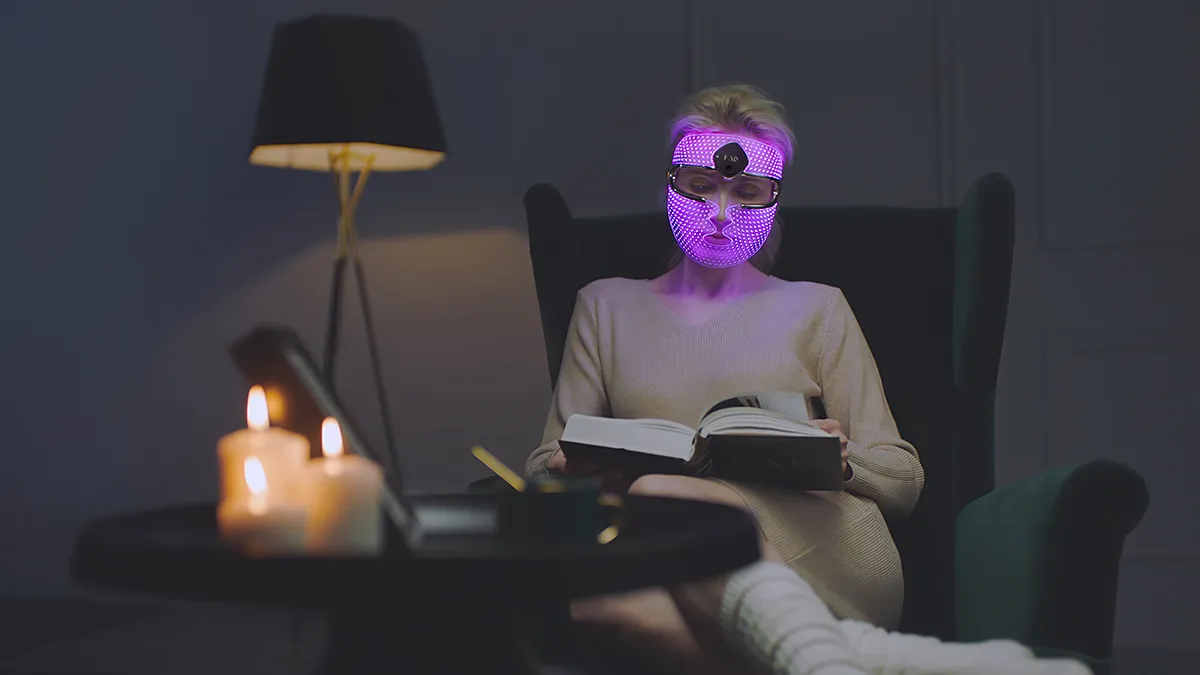 A woman reading on a cold night ina  cozy room with candles treating her face with FAQ 202 light therapy mask